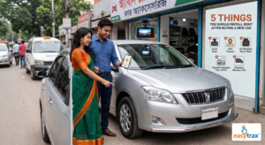 **A smiling young Bangladeshi couple, the woman in a green saree and the man in a blue shirt, stand by their new silver car outside a Dhaka car accessories shop. The man holds up his phone showing a live GPS tracking map of the vehicle. A large poster titled '5 THINGS YOU SHOULD INSTALL RIGHT AFTER BUYING A NEW CAR' lists five essential aftermarket products: Dash Cam, GPS Tracker, All-Weather Floor Mats, Paint Protection Film (PPF), and Premium Window Tint. The car has 'Dhaka Metro GA' plates, and a dash cam is visible mounted on the windshield. The background is a typical Dhaka street.**